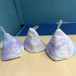 May include: Three fabric pyramid-shaped sachets with white loop handles. The sachets feature a watercolor design in shades of blue and purple, with gold geometric lines and celestial sun and moon designs.