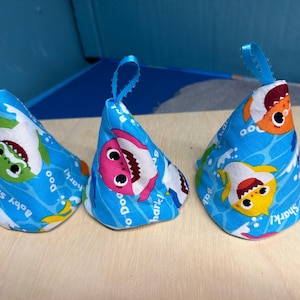 May include: Three blue fabric party hats with a Baby Shark theme. Each hat features a different shark character in bright colors, with the words "Baby Shark" and "Doo Doo" printed on the fabric. Each hat has a blue ribbon loop at the top.