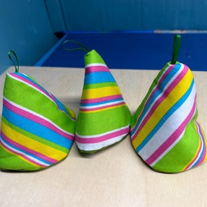 May include: Three cone-shaped fabric party hats with green, pink, yellow, and blue diagonal stripes. Each hat has a green loop at the top. The hats are on a light wood surface.