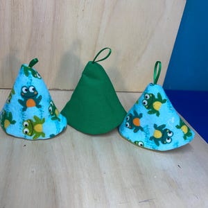 May include: Three cone-shaped fabric items. Two are light blue with a frog pattern, and one is solid green. Each has a green ribbon loop at the top. The items are displayed on a light wood surface.