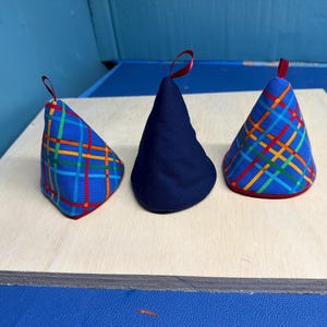 May include: Three fabric items in a cone shape. Two are blue with a plaid pattern in red, yellow, and green. The third is solid navy blue. Each has a red loop at the top. The items are on a light wood surface.