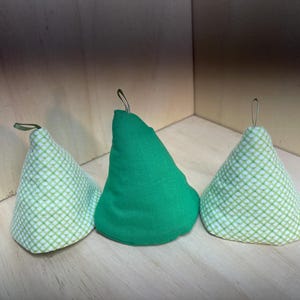 May include: Three fabric Christmas tree ornaments. Two are white with a green grid pattern, and one is solid green. Each has a small loop for hanging. The ornaments are cone-shaped.
