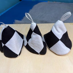 May include: Three black and white checkered fabric objects with white ribbon loops. The objects are cone-shaped and rest on a light wood surface. The pattern is a classic checkerboard design.