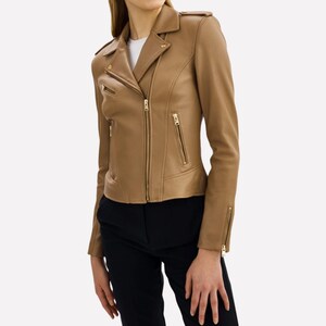 May include: A tan leather jacket with a classic biker style. The jacket features a notched collar, asymmetrical zipper closure, and multiple zippered pockets. The sleeves have zippered cuffs, and the jacket is worn over a white top and black pants.