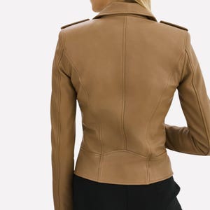 May include: Tan leather jacket with a tailored fit. The jacket features a classic collar, shoulder epaulets, and a fitted waist. The back view showcases the jacket's seams and design details. The model is wearing black pants.