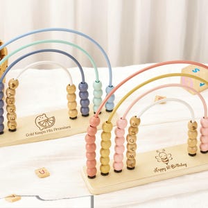 May include: Two wooden abacuses with rainbow-colored arches and wooden beads. One has the name "Jerry" on the beads, the other says "Abacus". The base of each has an image and text. A wooden block and stuffed animals are in the background.