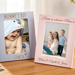 Puede incluir: Dos marcos de fotos de madera, uno azul claro y otro rosa, cada uno con una foto familiar. El marco azul claro dice "MUMMY'S BOY" y "EDWIN" con un corazón. El marco rosa dice "Home is wherever Mom is" y "Mom & Elspeth & James".