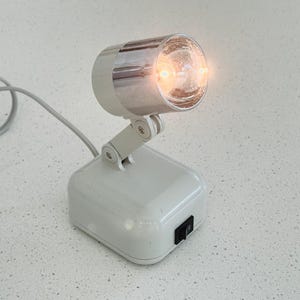 May include: A small white desk lamp with a chrome-finished cylindrical head and an adjustable hinged arm. The base is square with a black rocker power switch on the front. A light gray power cord extends from the back of the unit, and the lamp is illuminated with a warm glow.
