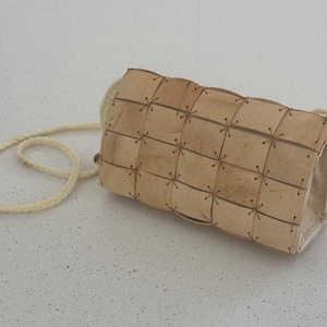 May include: A rectangular beige handbag constructed from a grid of small, square, light-colored panels joined together with thin, dark brown cord stitching. The bag features a braided, off-white shoulder strap attached to the sides.