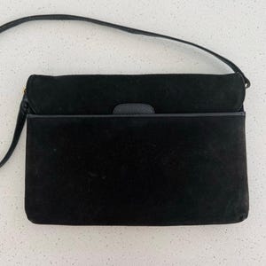 May include: A black suede shoulder bag featuring a rectangular silhouette, a fold-over flap closure, and a thin black shoulder strap. The bag includes a small, textured black tab detail on the front flap and smooth black leather trim along the edge.