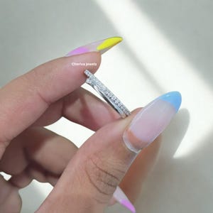 Puede incluir: Un anillo plateado con un diseño único, sostenido entre los dedos. El anillo presenta una fila de pequeñas piedras claras y una forma angular distintiva. El texto "Cheriva jewels" es visible.