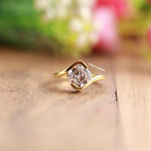 Puede incluir: Un anillo de oro con una gema cuadrada y transparente. El anillo tiene un diseño curvo único. El texto "Cheriva jewels" es visible en el anillo. El anillo está sobre un fondo borroso de flores.