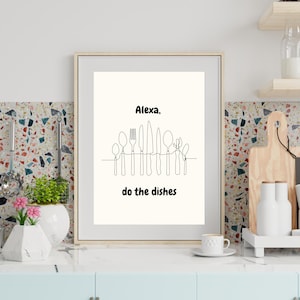 May include: Framed art print featuring a minimalist line drawing of cutlery. The text "Alexa, do the dishes" is above the utensils. The artwork is in a light-colored frame, set against a kitchen backdrop with a blue cabinet and a white countertop.