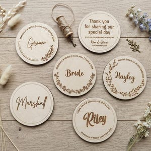May include: Six circular light-coloured wooden discs featuring laser-engraved text and floral motifs. The discs display names such as Groom, Bride, Marshall, Hayley, and Riley, alongside wedding dates and the text Thank you for sharing our special day.