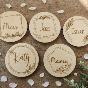 May include: Five circular wooden place cards, each engraved with a name: Marie, Jase, Oscar, Katy, and Marie. The cards have different geometric frames and floral or leaf designs. They are displayed on a wooden surface with scattered petals.