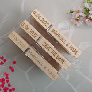 May include: Three light-coloured wooden clothespins with metal springs. Two pins are engraved with the text Marshall & Marie and the date 01.06.2022. The middle pin is engraved with the text Save The Date and the date 01.06.2022.