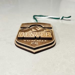 May include: Wooden Denver Summit FC ornament. The ornament is shield-shaped with a mountain range design above the word "DENVER". The text "SUMMIT FC" is below. It has a green and white string for hanging.