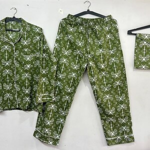 May include: Green pajama set with a white palm tree pattern. The set includes a button-down shirt, pants with a drawstring waist, and a matching eye mask. The pajamas are hanging on black hangers against a white background.
