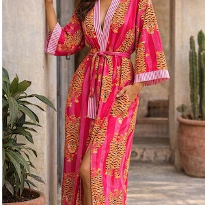 May include: A long, flowing pink robe with a tiger stripe pattern. The robe has a V-neck, wide sleeves, and a matching belt. The sleeves and trim feature a white and pink striped design. The robe is worn open, revealing a slit up the leg.