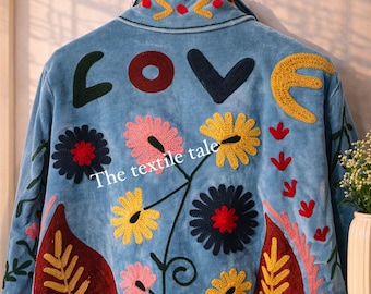 May include: Light blue jacket with embroidered floral and leaf designs. The word "LOVE" is embroidered on the back, along with the text "The textile tale". The jacket is hanging on a wooden hanger.