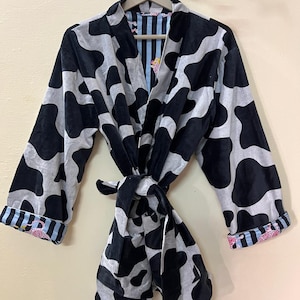 May include: A black and white patterned robe with a tie closure. The robe features a cow print design and striped and floral patterned cuffs. The robe is hanging on a wooden hanger.