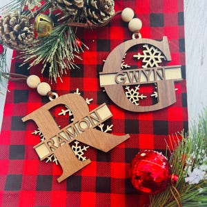 May include: Two wooden Christmas ornaments with personalized names, "GWYN" and "RAMON", are displayed on a red and black plaid background. The ornaments are in the shape of the first letter of the name, with snowflake cutouts. Festive holiday decor surrounds the ornaments.