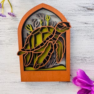 May include: A layered wooden art piece featuring a sea turtle design. The turtle is depicted in shades of green and black, set against a yellow background. The frame is a warm orange color, with coral and seaweed accents.