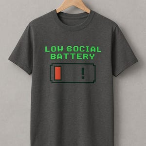 Può includere: T-shirt grigio scuro con la scritta "LOW SOCIAL BATTERY" in verde a caratteri pixelati sopra una grafica a batteria pixelata. La grafica della batteria mostra una carica bassa con una barra rossa e un punto esclamativo.