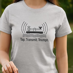 Può includere: T-shirt grigio chiaro con grafica nera di un tasto telegrafico, onde sonore e la scritta "Tap. Transmit. Triumph." Un tasto telegrafico vintage è visibile in primo piano.