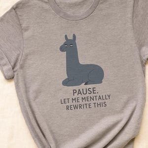 Può includere: T-shirt grigio con un'illustrazione di un lama blu e la scritta "PAUSE. LET ME MENTALLY REWRITE THIS". La maglietta ha un girocollo ed è realizzata in tessuto morbido.