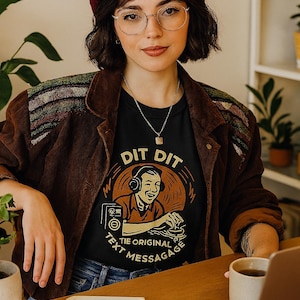 Può includere: Una persona indossa una maglietta nera con la scritta "DIT DIT TEXT ORIGINAL MESSAGE" e una grafica di una persona con le cuffie. La persona indossa un basco bordeaux e occhiali. È seduta a una scrivania di legno con un laptop e una tazza di caffè.