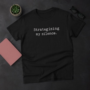 Può includere: T-shirt nero con la scritta bianca "Strategizing my silence". Sono visibili anche una piccola pianta grassa in vaso, un quaderno rosa e un paio di occhiali neri. La maglietta è in cotone.