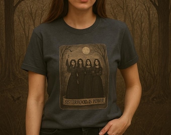 Op de afbeelding: Donkergrijs t-shirt met de tekst "SISTERHOOD IS POWER" en een afbeelding van vier vrouwen in lange jurken. De achtergrond is een boslandschap met een volle maan en een vleermuis. De persoon draagt een zwarte jeans.