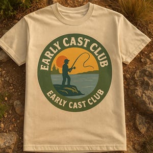 Può includere: T-shirt color crema con grafica circolare con la scritta "EARLY CAST CLUB". La grafica raffigura un pescatore che lancia la lenza contro un tramonto. La maglietta è realizzata in materiale morbido, adatta alle attività all'aperto.