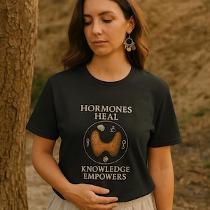 May include: A black t-shirt with the text "HORMONES HEAL" and "KNOWLEDGE EMPOWERS" in a light brown font. The shirt features a graphic of a thyroid gland. The person is wearing beige pants and gold earrings.