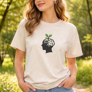 Può includere: T-shirt beige a maniche corte con silhouette nera di testa, design del cervello e foglie verdi. La modella indossa jeans blu. Lo sfondo è una scena all'aperto sfocata con alberi e vegetazione.