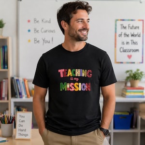Può includere: T-shirt nero con la scritta "TEACHING is my MISSION" in lettere colorate e decorate. La persona sorride in un'aula, con scaffali e poster motivazionali sullo sfondo.