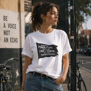 Può includere: Una t-shirt bianca a maniche corte con una grafica a bandiera nera sul petto. La grafica contiene il testo: When the power of love beats the love of power the world knows peace. La maglietta ha un girocollo e maniche arrotolate.