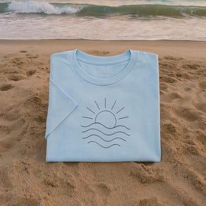 Puede incluir: Camiseta azul claro con un dise&ntilde;o gr&aacute;fico negro minimalista de un sol sobre olas. La camiseta est&aacute; doblada sobre una playa de arena, con el mar y las olas al fondo.
