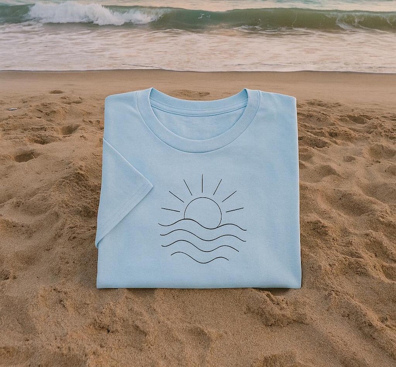Puede incluir: Camiseta azul claro con un dise&ntilde;o gr&aacute;fico negro minimalista de un sol sobre olas. La camiseta est&aacute; doblada sobre una playa de arena, con el mar y las olas al fondo.