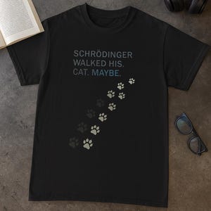 Könnte beinhalten: Schwarzes T-Shirt mit dem Text "SCHRÖDINGER WALKED HIS. CAT. MAYBE." in Blau und Grau. Eine Reihe von Pfotenabdrücken in verschiedenen Grau- und Weißtönen zieht sich über das Shirt und erzeugt ein visuelles Design.