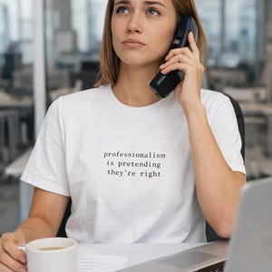 Può includere: Una persona indossa una maglietta bianca con la scritta "professionalism is pretending they're right" e parla al telefono. Una tazza bianca e un laptop sono sulla scrivania. L'ambientazione sembra essere un ufficio.