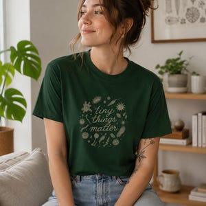 Könnte beinhalten: Dunkelgrünes T-Shirt mit dem Text "tiny things matter" in einem kreisförmigen Design. Das Design enthält verschiedene kleine Illustrationen. Das Shirt wird mit einer blauen Jeans kombiniert.