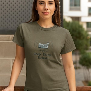 May include: Olive green t-shirt with a graphic of a radio and the text "Beep. Think. Repeat." The shirt is made of a soft material and has short sleeves. The model is wearing blue jeans.