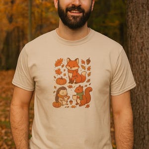 Può includere: T-shirt beige con grafica a tema autunnale. Il design include una volpe, un riccio e uno scoiattolo, ognuno con una tazza, circondati da zucche, ghiande e foglie. La maglietta è realizzata in materiale morbido.