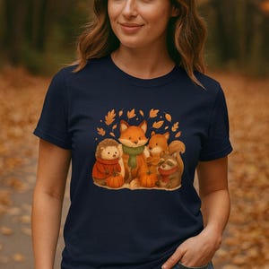 Può includere: T-shirt blu navy con un'illustrazione ad acquerello di una volpe, uno scoiattolo, un procione e un riccio. Gli animali sono circondati da foglie autunnali e zucche. La maglietta ha un girocollo.