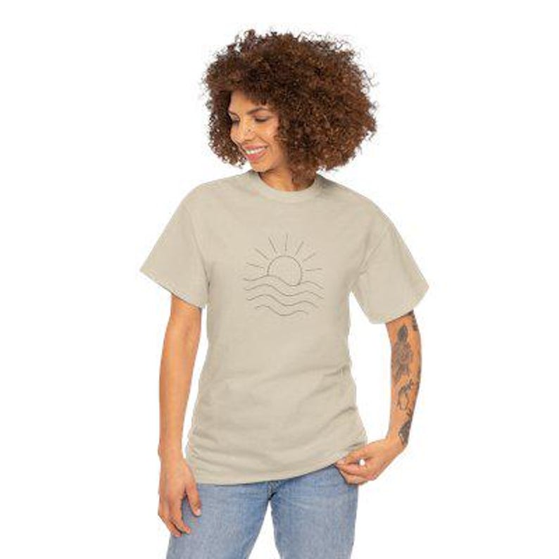 Puede incluir: Una camiseta beige con un dibujo lineal minimalista de un sol sobre l&iacute;neas onduladas. La camiseta de manga corta la lleva una persona con vaqueros azules. El dise&ntilde;o est&aacute; centrado en la parte delantera de la camiseta.