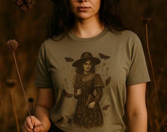 Op de afbeelding: Olijfgroen t-shirt met een sepia-getinte illustratie van een vrouw met een hoed, die een boek vasthoudt, omringd door vlinders. De afbeelding heeft een vintage esthetiek. De persoon draagt een bruine hoed en een zwarte jeans.
