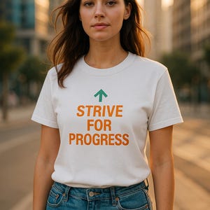 Può includere: Una maglietta bianca con le parole "STRIVE FOR PROGRESS" in arancione e una freccia verde rivolta verso l'alto. La persona indossa jeans blu. Lo sfondo è una scena urbana sfocata.