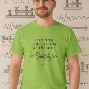 Può includere: Una maglietta verde lime con la scritta "LISTEN TO THE RHYTHM OF THE DOTS" e una grafica di un tasto Morse. Lo sfondo presenta grafici del codice Morse.
